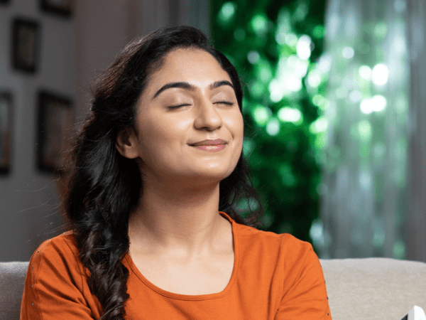 A relaxed woman in an orange shirt sits comfortably with her eyes closed, enjoying the fresh air inside her home. This image represents the importance of Indoor Air Quality for creating a healthy and comfortable living environment.